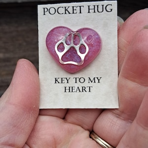May include: A heart-shaped pocket hug with a silver paw print design. The heart is a mix of pink and purple with the words "POCKET HUG" and "KEY TO MY HEART" printed on the card.
