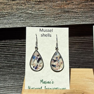 May include: Teardrop-shaped earrings with a silver-colored frame, filled with pieces of mussel shells. The earrings hang from silver hooks. The card behind the earrings reads "Mussel shells" and "Maine's Natural Inspirations".