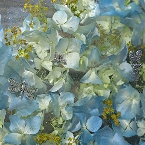 May include: A close-up of a resin art piece featuring blue and white hydrangea flowers and silver dragonfly accents. The flowers are pressed and preserved in the resin, creating a unique and beautiful piece of art.