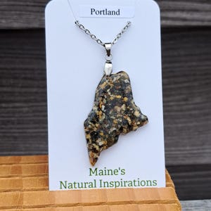 May include: A pendant necklace featuring a pendant in the shape of the state of Maine. The pendant is made of a speckled, multi-colored material and hangs from a silver chain. The card behind the pendant reads "Portland" and "Maine's Natural Inspirations."