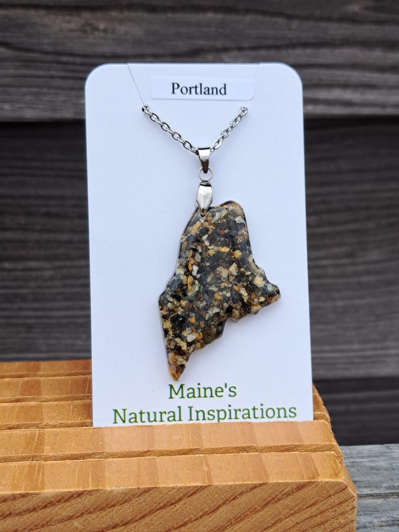 May include: A pendant necklace featuring a pendant in the shape of the state of Maine. The pendant is made of a speckled, multi-colored material and hangs from a silver chain. The card behind the pendant reads "Portland" and "Maine's Natural Inspirations."