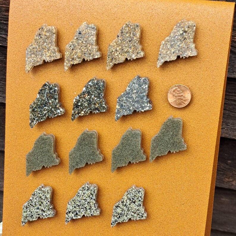 May include: A collection of 15 small, decorative, state-shaped ornaments. The ornaments are made of a clear material filled with sand and glitter. The ornaments are arranged in three rows of five. The ornaments are all in the shape of the state of Maine.