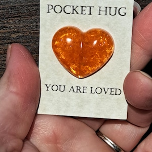 May include: A small card with the words "POCKET HUG" and "YOU ARE LOVED" printed on it. A heart-shaped, orange-colored stone is attached to the card. The stone has a textured appearance and is held in a person's hand.