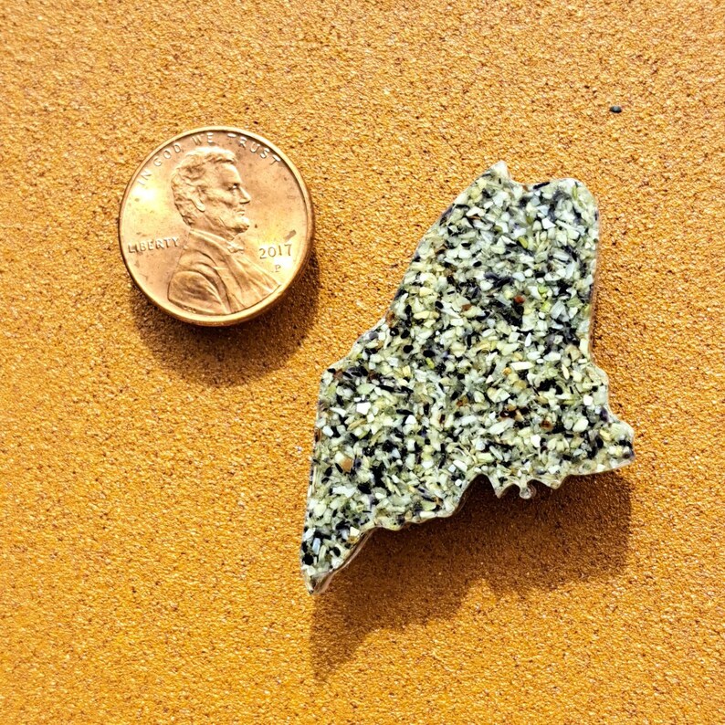 May include: A small, green and black, state-shaped, mosaic tile of Maine. The tile is about the size of a US penny, which is shown in the image for scale.
