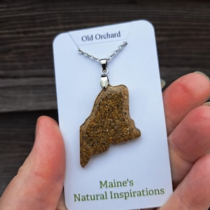 May include: A pendant necklace featuring a pendant in the shape of the state of Maine. The pendant is a speckled brown color and is attached to a silver chain. The card reads "Old Orchard" and "Maine's Natural Inspirations."