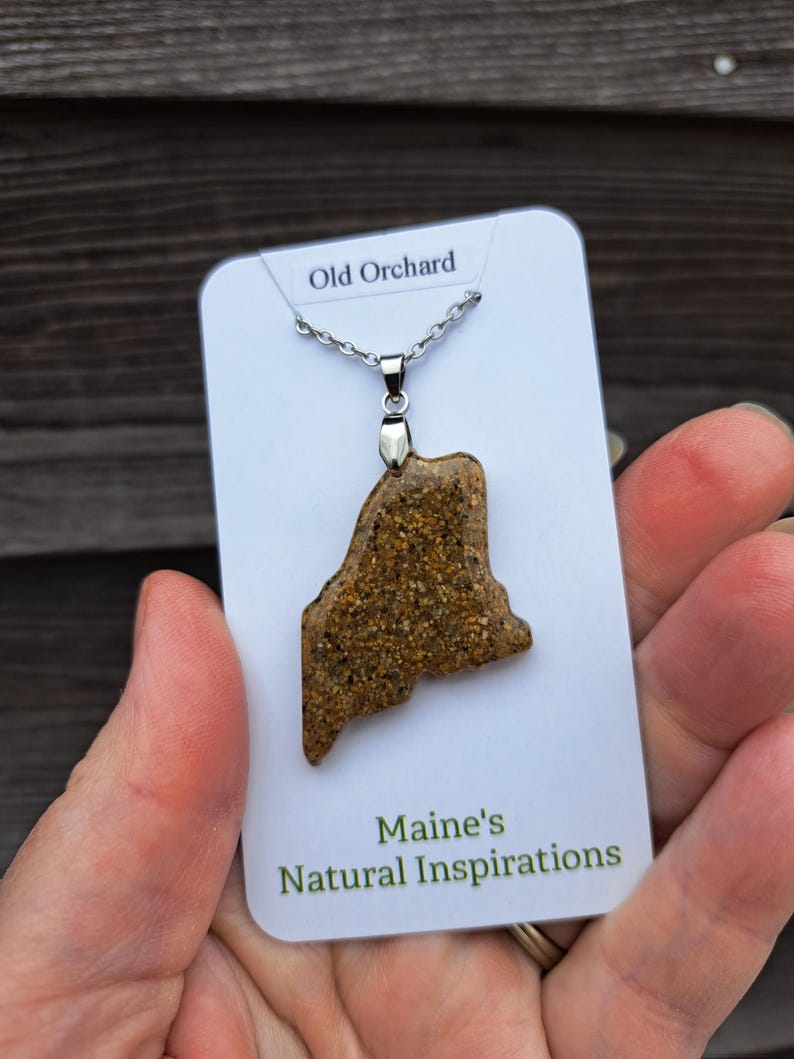 May include: A pendant necklace featuring a pendant in the shape of the state of Maine. The pendant is a speckled brown color and is attached to a silver chain. The card reads "Old Orchard" and "Maine's Natural Inspirations."