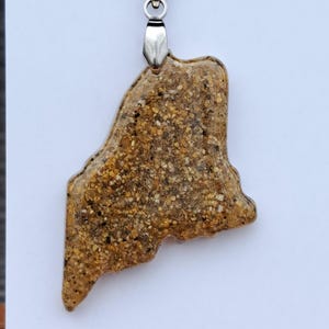 May include: A pendant necklace featuring a pendant in the shape of the state of Maine. The pendant is made of a sand-like material with a mix of tan, brown, and black grains. The necklace has a silver-colored chain and clasp. The text "Maine's" is visible.
