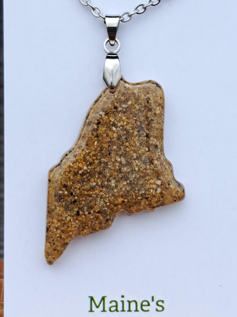 May include: A pendant necklace featuring a pendant in the shape of the state of Maine. The pendant is made of a sand-like material with a mix of tan, brown, and black grains. The necklace has a silver-colored chain and clasp. The text "Maine's" is visible.