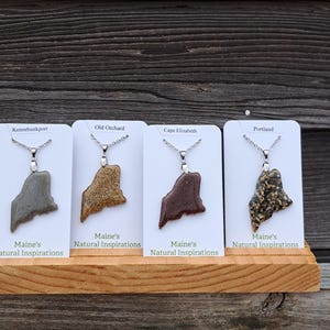 May include: Four necklaces with pendants shaped like the state of Maine, each on a white card. The pendants are in shades of gray, brown, and black, with silver chains. The cards are labeled with Maine towns and "Maine's Natural Inspirations".