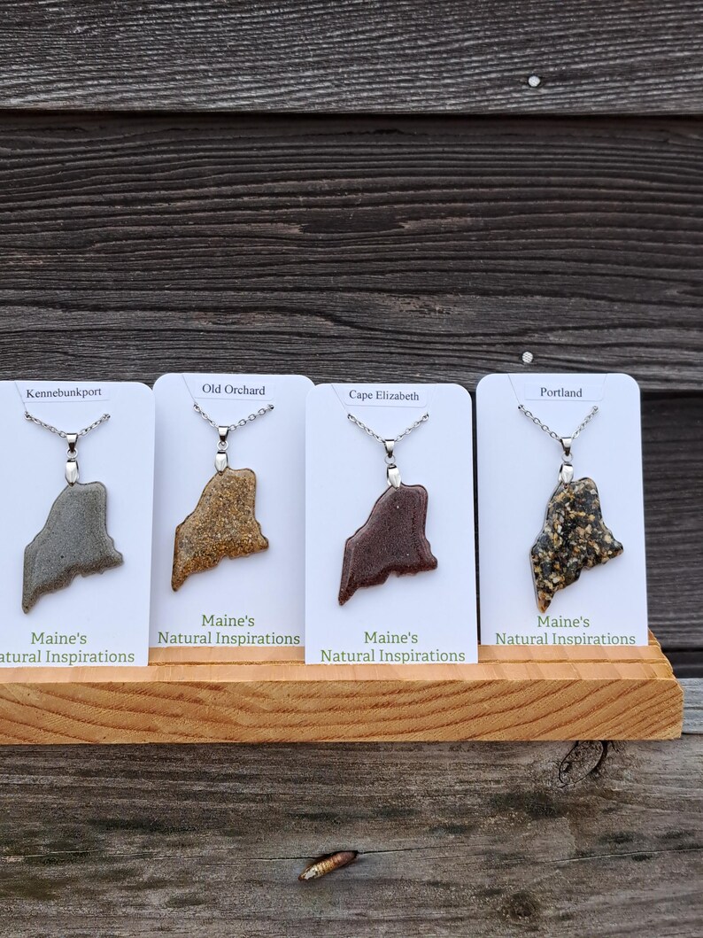May include: Four necklaces with pendants shaped like the state of Maine, each on a white card. The pendants are in shades of gray, brown, and black, with silver chains. The cards are labeled with Maine towns and "Maine's Natural Inspirations".