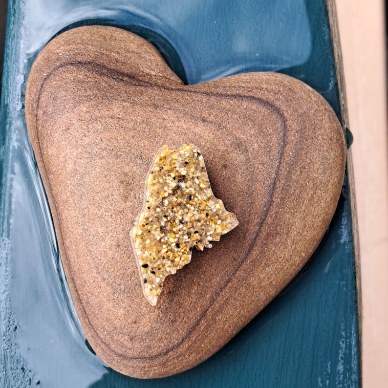 May include: A heart-shaped stone with a golden glitter outline of the state of Maine. The stone is resting on a teal blue surface.