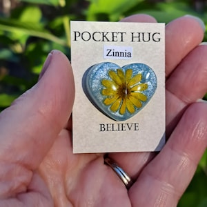 May include: A heart-shaped Pocket Hug with a yellow zinnia flower encased in clear resin with a blue background. The card reads "POCKET HUG" at the top, "Zinnia" in the middle, and "BELIEVE" at the bottom.