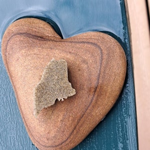 May include: A heart-shaped stone with a smaller stone shaped like the state of Maine on top. The heart-shaped stone is brown and the smaller stone is a lighter brown.