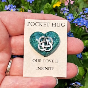 May include: A teal heart-shaped Pocket Hug with a silver Celtic knot design, held in a hand. The card reads "POCKET HUG" and "OUR LOVE IS INFINITE". The background features blue flowers and green foliage.