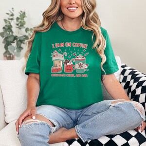 Puede incluir: Una camiseta verde con el texto "I RUN ON COFFEE, CHRISTMAS CHEER, AND BATS" en rojo y blanco. La camiseta presenta tres diseños de tazas de café con decoraciones navideñas. La camiseta es de cuello redondo y está hecha de un material suave.