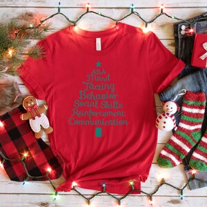 May include: Red t-shirt with a teal green Christmas tree design. The tree is decorated with the words "ABA", "Mand", "Teaching", "Behavior", "Social Skills", "Reinforcement", and "Communication".