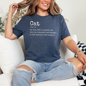 Peut inclure: Un t-shirt bleu marine avec du texte blanc qui dit "Cat I kat I n. the furry little sociopath who rules my household and manages to look innocent while doing so."