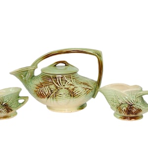 May include: A green and brown ceramic teapot and two matching creamer and sugar bowls. The teapot has a handle and a lid. The creamer and sugar bowls have a single handle. All three pieces feature a pine cone design.