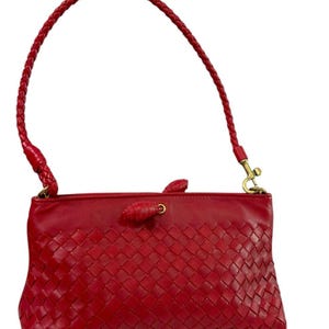 May include: A red leather handbag with a woven pattern on the front. The bag has a braided handle and a gold-colored clasp. The bag is rectangular and has a small, decorative knot detail.