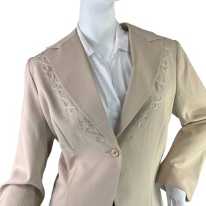 Puede incluir: Una chaqueta beige con un cierre de botón único y un diseño de cuentas decorativas en la solapa.