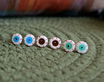 May include: Six small, round earrings with a white base, each featuring a different colored iris: blue, brown, and green. The earrings are arranged in a row on a textured green surface. A plaid pillow is in the background.