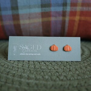 May include: A pair of orange pumpkin-shaped polymer clay stud earrings. The earrings are displayed on a light green card with the text "SAGED siren" and "polymer clay earrings and studs". The background includes a plaid pillow and a green woven mat.