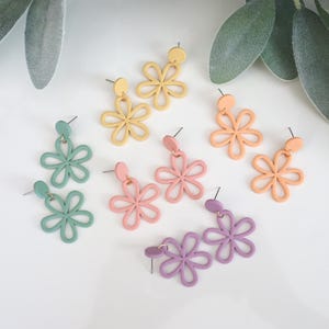 May include: A collection of colorful flower-shaped earrings in pastel shades of green, yellow, pink, peach, and purple. The earrings are displayed on a white surface with green foliage in the background. The earrings are made of a lightweight material.