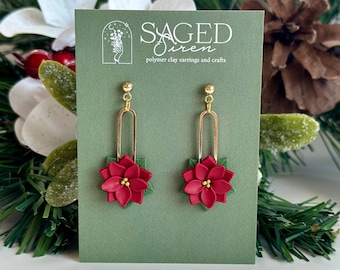 Op de afbeelding: Een paar goudkleurige oorhangers met rode poinsettia bloemmotieven en groene bladeren. De oorbellen worden gepresenteerd op een groene kaart met de tekst "Saged Siren polymer clay earrings and crafts". De achtergrond bevat groen en witte bloemen.