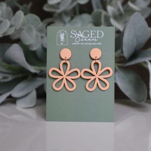May include: A pair of peach-colored polymer clay earrings in a floral design. The earrings are displayed on a green card with the text "SAGED Siren polymer clay earrings and crafts". The background features green foliage.