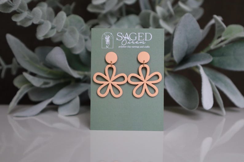 May include: A pair of peach-colored polymer clay earrings in a floral design. The earrings are displayed on a green card with the text "SAGED Siren polymer clay earrings and crafts". The background features green foliage.