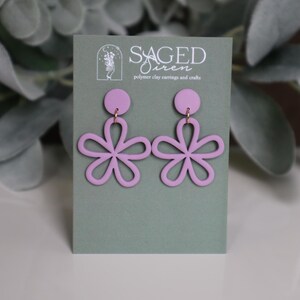 May include: A pair of light purple flower-shaped earrings. The earrings are made of polymer clay and have a circular stud at the top. The flower design has five petals. The earrings are displayed on a green card with the text "SAGED Siren polymer clay earrings and crafts."