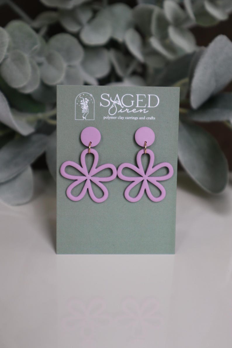 May include: A pair of light purple flower-shaped earrings. The earrings are made of polymer clay and have a circular stud at the top. The flower design has five petals. The earrings are displayed on a green card with the text "SAGED Siren polymer clay earrings and crafts."