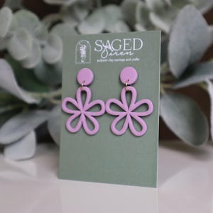 May include: A pair of light purple flower-shaped earrings with gold-colored hooks are displayed on a green card. The card has the text "SAGED Siren" and "polymer clay earrings and crafts". The earrings are set against a blurred background of green foliage.
