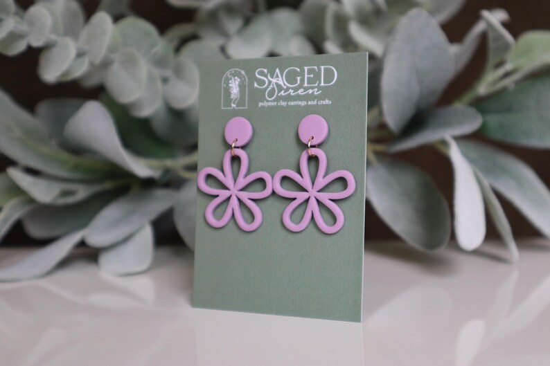 May include: A pair of light purple flower-shaped earrings with gold-colored hooks are displayed on a green card. The card has the text "SAGED Siren" and "polymer clay earrings and crafts". The earrings are set against a blurred background of green foliage.