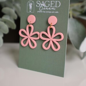 May include: A pair of light pink polymer clay earrings in a floral design. Each earring features a circular stud and a flower with five petals. The earrings are displayed on a green card with the text "Saged Siren" and "polymer clay earrings and crafts".