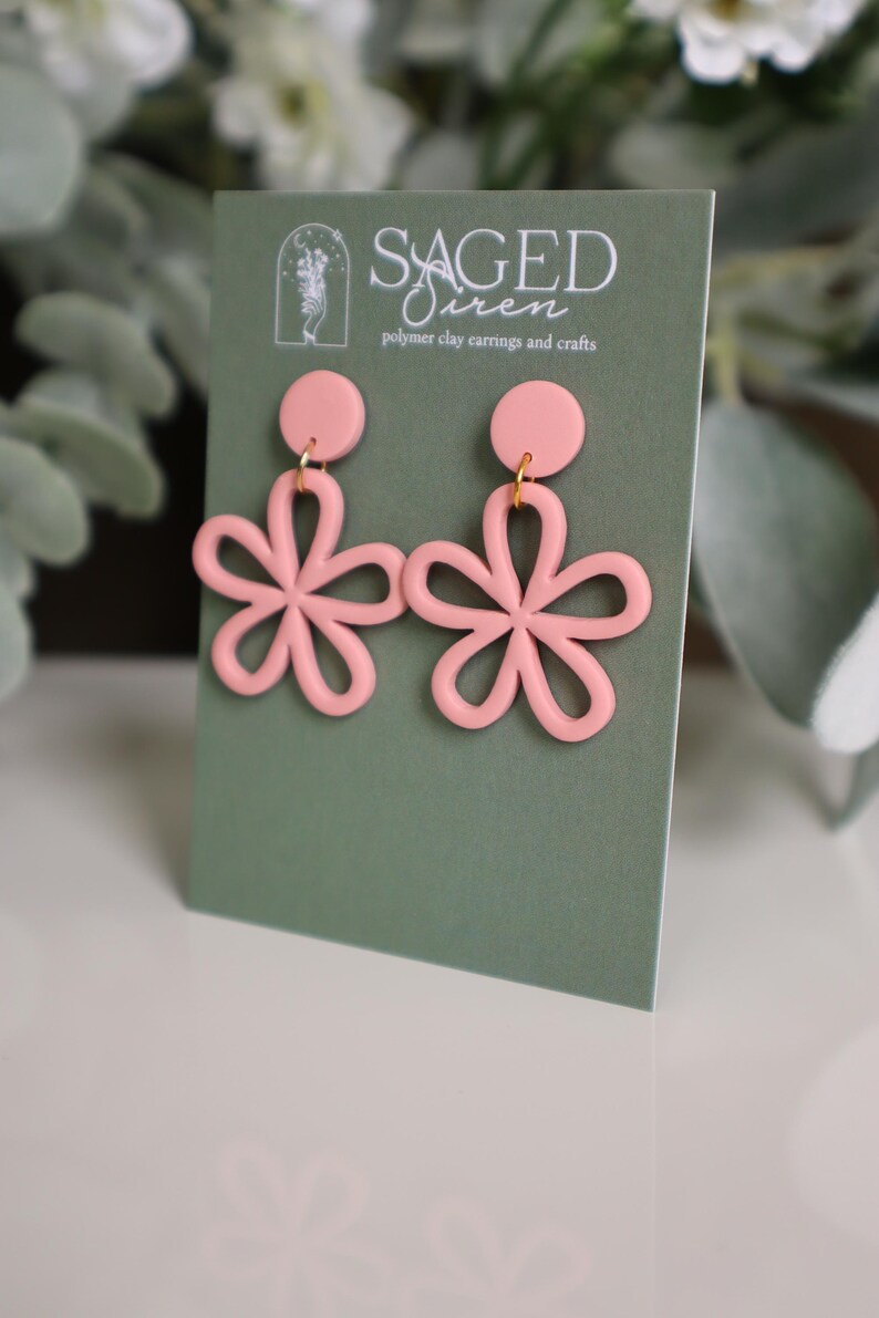 May include: A pair of light pink polymer clay earrings in a floral design. Each earring features a circular stud and a flower with five petals. The earrings are displayed on a green card with the text "Saged Siren" and "polymer clay earrings and crafts".