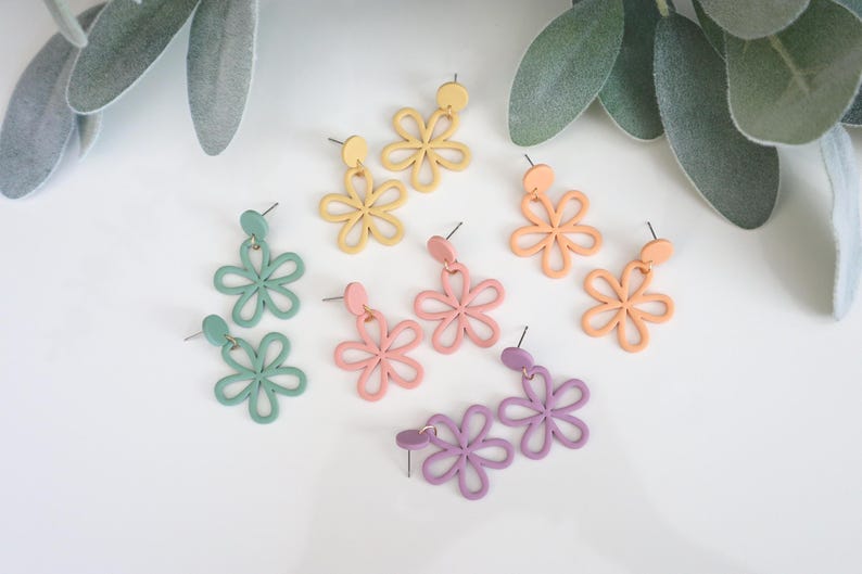 May include: A collection of colorful flower-shaped earrings in pastel shades of green, yellow, pink, peach, and purple. The earrings are displayed on a white surface with green foliage in the background. The earrings are made of a lightweight material.