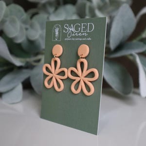 May include: A pair of peach-colored polymer clay earrings in a floral design. Each earring features a circular stud and a flower with six petals. The earrings are displayed on a green card with the text "SAGED Siren polymer clay earrings and crafts."