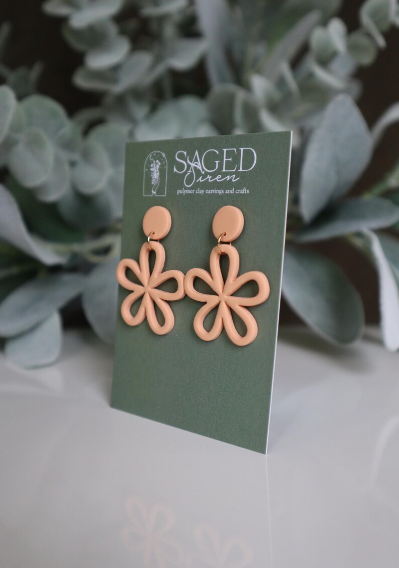 May include: A pair of peach-colored polymer clay earrings in a floral design. Each earring features a circular stud and a flower with six petals. The earrings are displayed on a green card with the text "SAGED Siren polymer clay earrings and crafts."