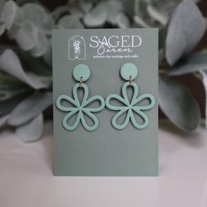 May include: Sage green polymer clay earrings in a floral design. The earrings are displayed on a sage green card with the text "Saged Siren polymer clay earrings and crafts". The earrings have a circular top and a flower shape.