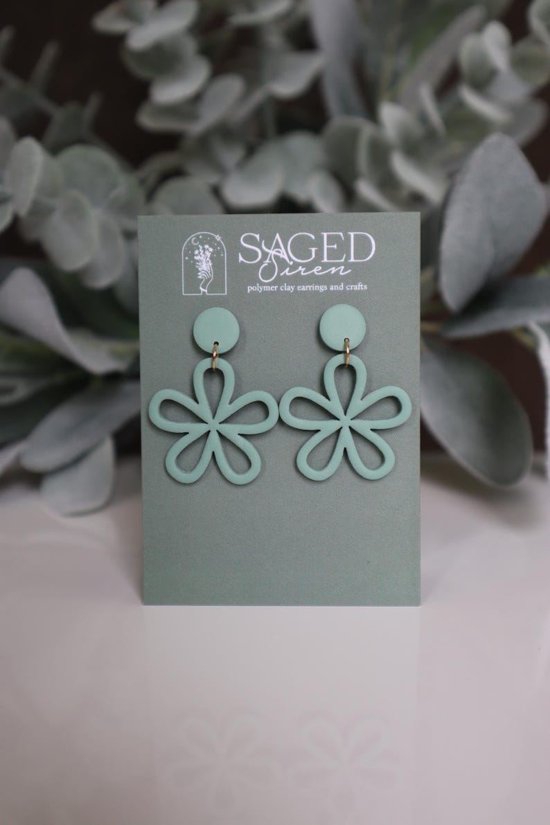 May include: Sage green polymer clay earrings in a floral design. The earrings are displayed on a sage green card with the text "Saged Siren polymer clay earrings and crafts". The earrings have a circular top and a flower shape.