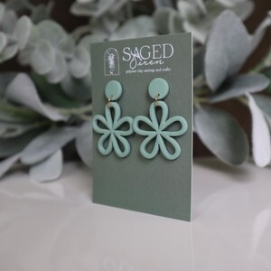 May include: Mint green flower-shaped polymer clay earrings with gold-colored hardware. The earrings are displayed on a sage green card with the text "SAGED SIREN" and the tagline "polymer clay earrings and crafts". The background features green foliage.