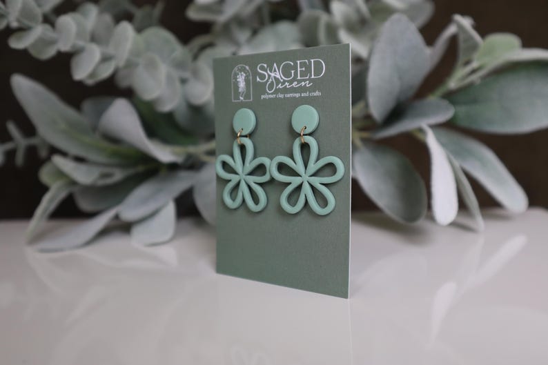 May include: Mint green flower-shaped polymer clay earrings with gold-colored hardware. The earrings are displayed on a sage green card with the text "SAGED SIREN" and the tagline "polymer clay earrings and crafts". The background features green foliage.