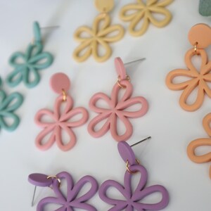 May include: A collection of colorful flower-shaped earrings in various pastel shades, including light blue, pink, yellow, peach, and purple. The earrings have a simple, open design and are displayed on a white surface.