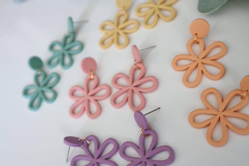 May include: A collection of colorful flower-shaped earrings in various pastel shades, including light blue, pink, yellow, peach, and purple. The earrings have a simple, open design and are displayed on a white surface.