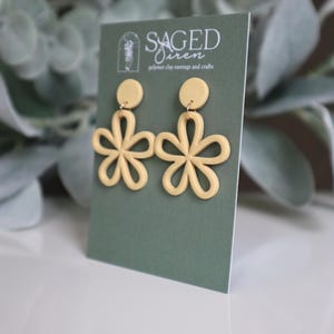 May include: A pair of light yellow flower-shaped polymer clay earrings. Each earring features a circular stud and a five-petal flower design. The earrings are displayed on a green card with the text "SAGED Siren polymer clay earrings and crafts".