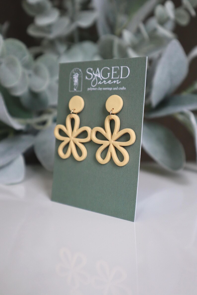 May include: A pair of light yellow flower-shaped polymer clay earrings. Each earring features a circular stud and a five-petal flower design. The earrings are displayed on a green card with the text "SAGED Siren polymer clay earrings and crafts".