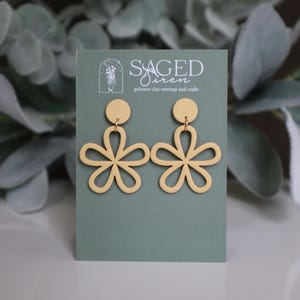 May include: A pair of pale yellow polymer clay earrings in a floral design. Each earring features a circular stud and a flower shape with five petals. The earrings are displayed on a green card with the text "SAGED Siren polymer clay earrings and crafts".