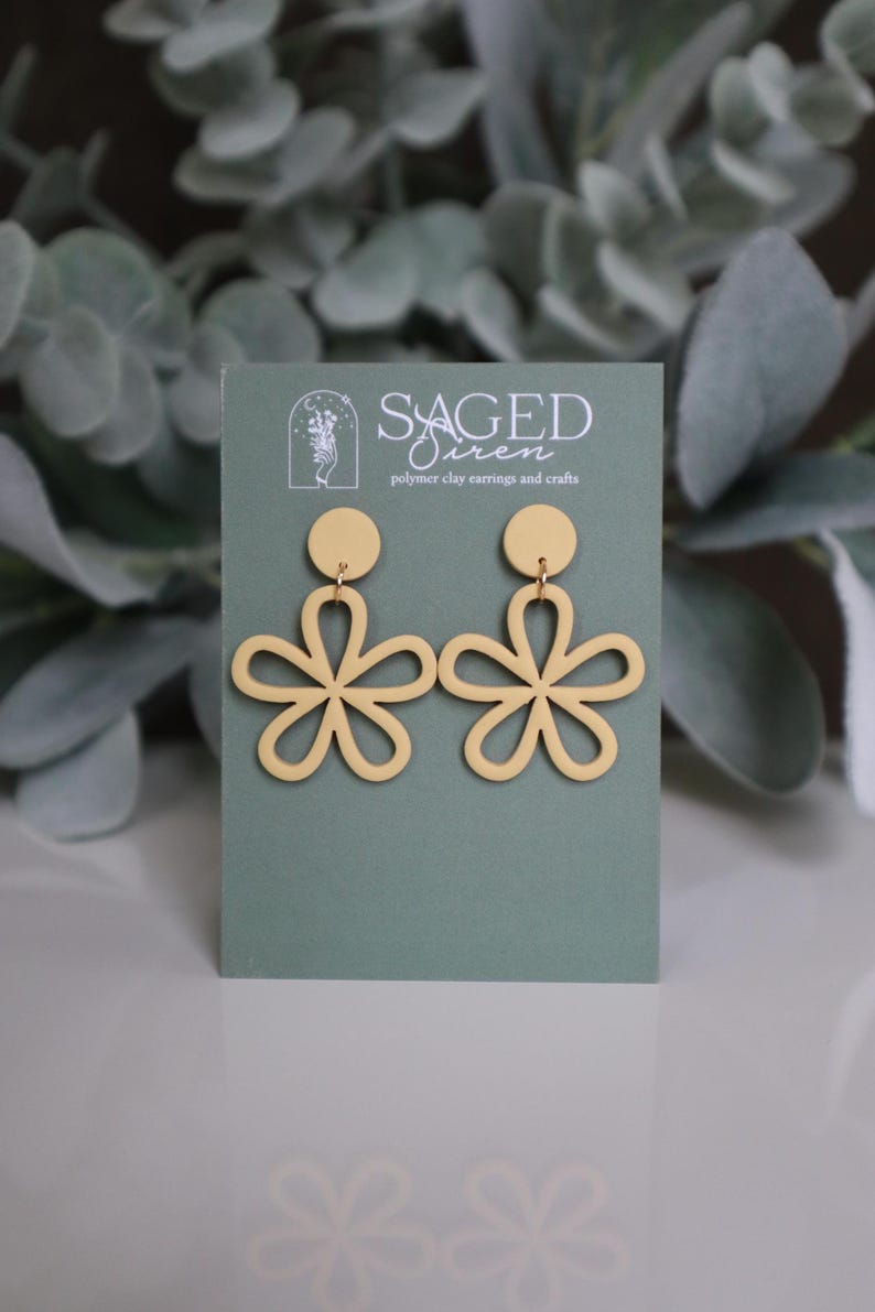 May include: A pair of pale yellow polymer clay earrings in a floral design. Each earring features a circular stud and a flower shape with five petals. The earrings are displayed on a green card with the text "SAGED Siren polymer clay earrings and crafts".