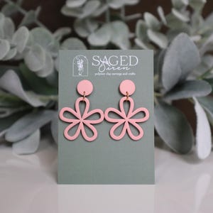 May include: Pink flower-shaped earrings with a circular top and a floral design. The earrings are displayed on a gray card with the text "SAGED Siren polymer clay earrings and crafts". The background features green foliage.